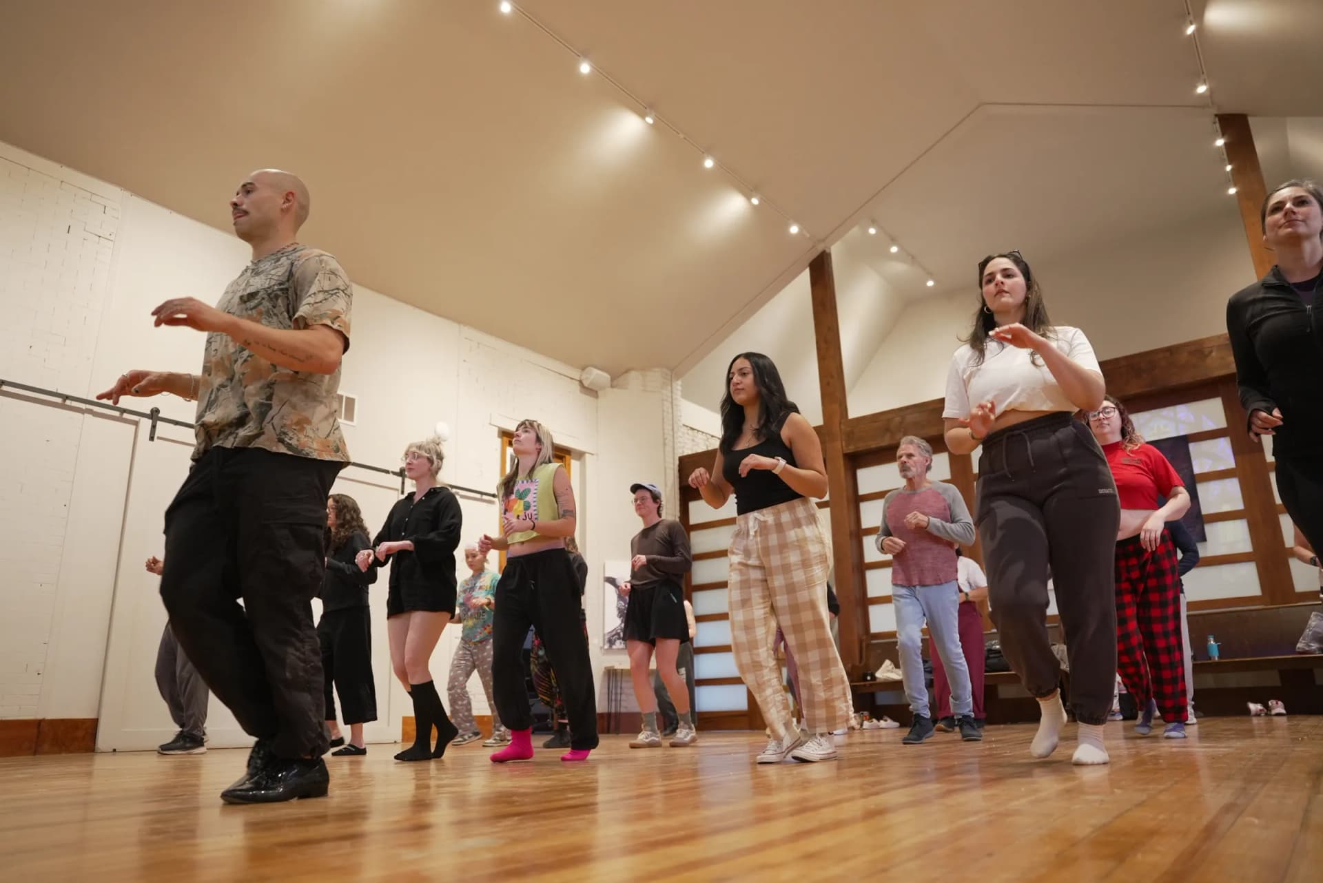 A dance class moves in step across the wood floor of the hall under strings of warm lights