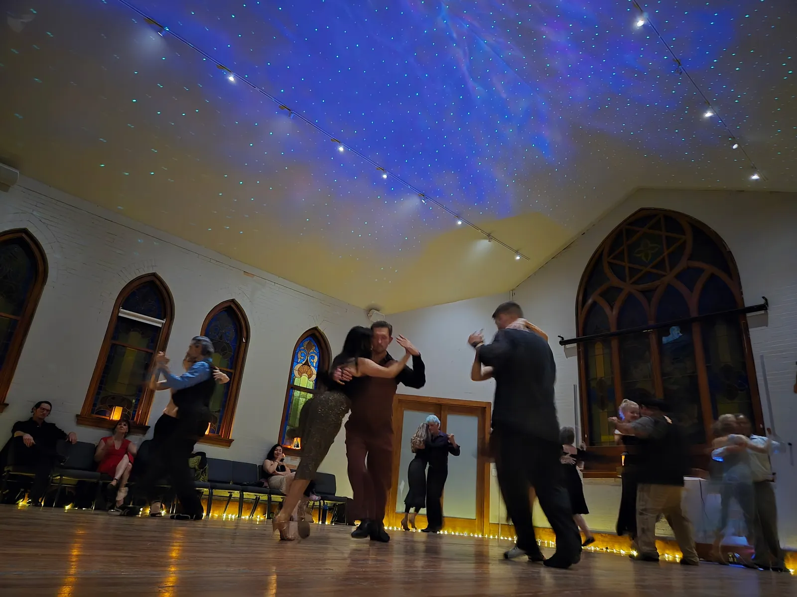 Couples dance beneath a projected night sky, the tall arched window glowing behind