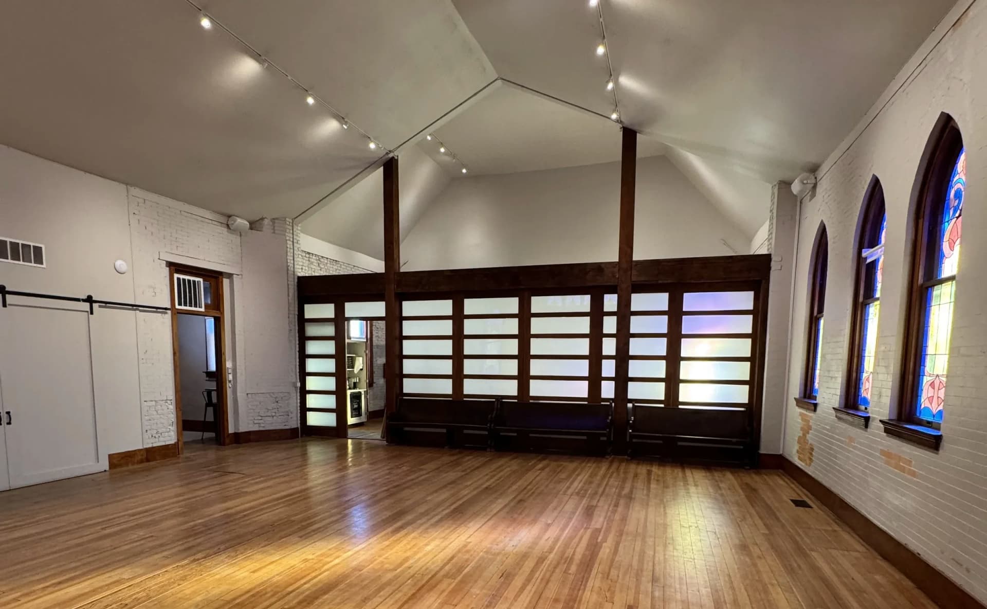 Sunlight streaks the long wood floor of the empty hall, facing the wood-framed partition wall and arched windows